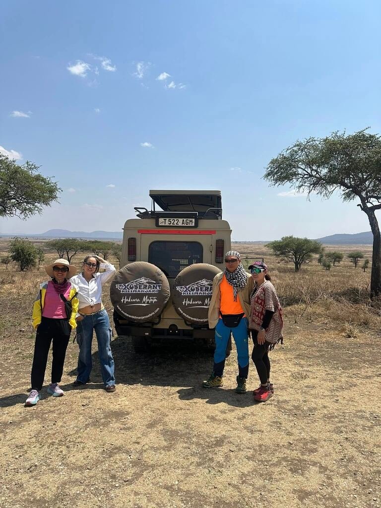 Four travellers beside an Exuberant Kilimanjaro safari vehicle in an open savannah landscape.