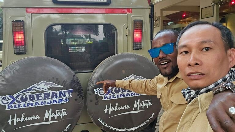 Two people beside an Exuberant Kilimanjaro safari vehicle with branded spare-wheel covers.